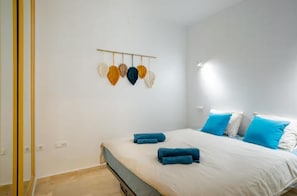 Une chambre à coucher avec un lit, des serviettes bleues, du linge de lit blanc et un luminaire fixé au mur.