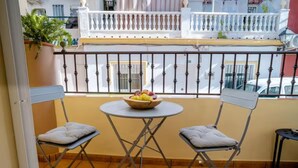 A balcony with a table and two chairs, a potted plant, and a view of a white building with balconies.