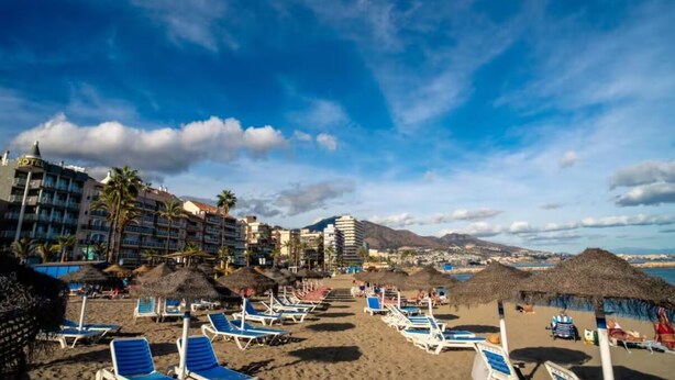 A beach with sun loungers, palm trees, and a hotel in the background.