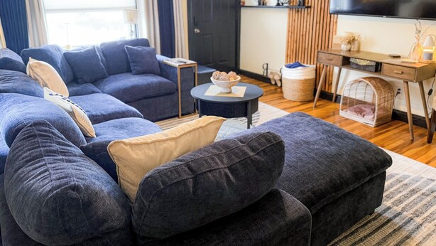 Een woonkamer met een hoekbank, een salontafel, een televisie, een bureau en een huisdierenbed.