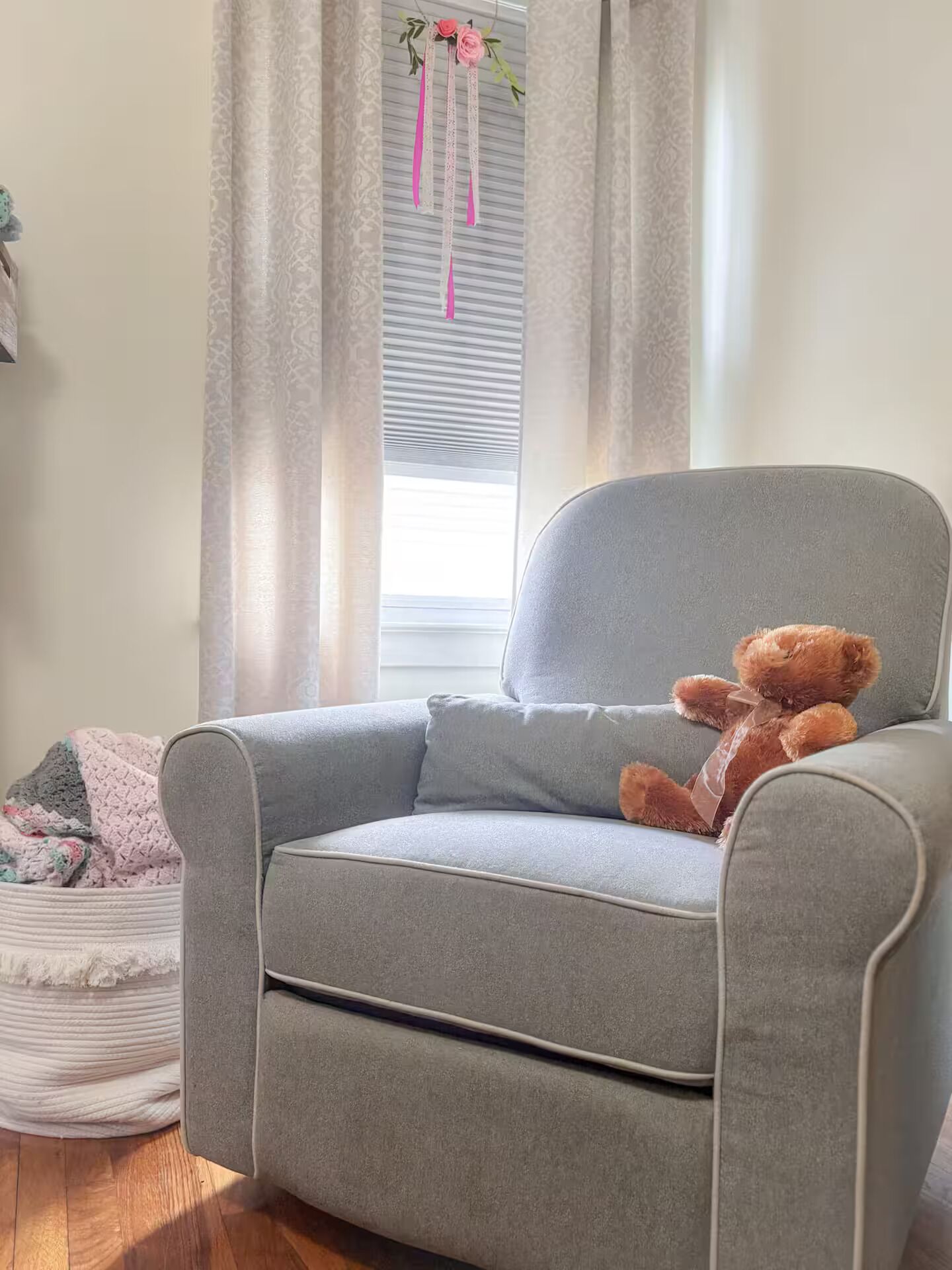 Ein grauer Sessel mit einem Teddybär, ein Fenster mit Jalousien und einem gemusterten Vorhang.