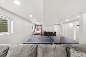 A room with two ping pong tables, a foosball table, and a basketball hoop.
