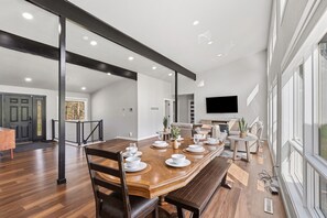 A spacious dining area with a long wooden table set for six, a modern kitchen in the background, and a large window providing natural light.