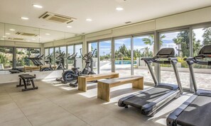 Ein Fitnessraum mit Laufbändern, Ergometer und Bänken, große Fenster mit Blick auf einen Pool und Grünflächen.