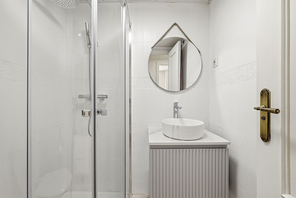 Un bany amb plat de dutxa de vidre, un mirall rodó i un lavabo blanc amb taulell de marbre.