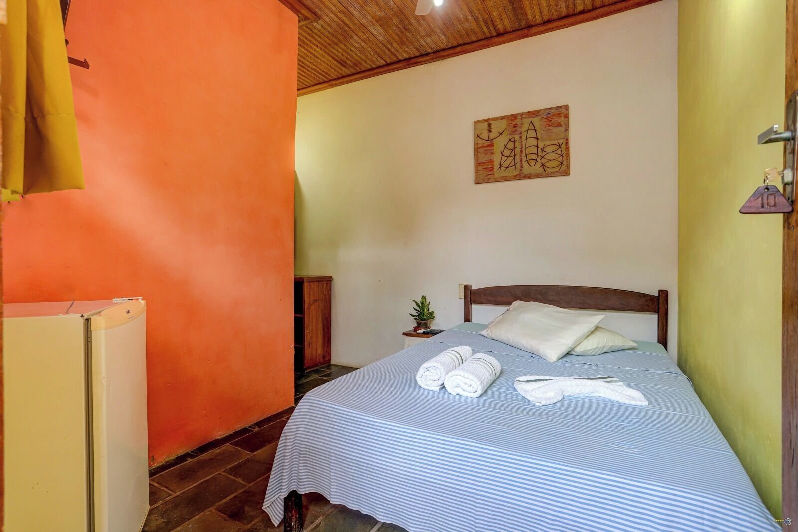 A single bed with a striped bedspread, two rolled towels, a small potted plant, and a wooden headboard.