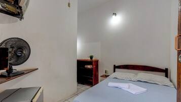 Un dormitorio con una cama, un escritorio, una silla, una mesita y un ventilador de pared, además de una puerta.