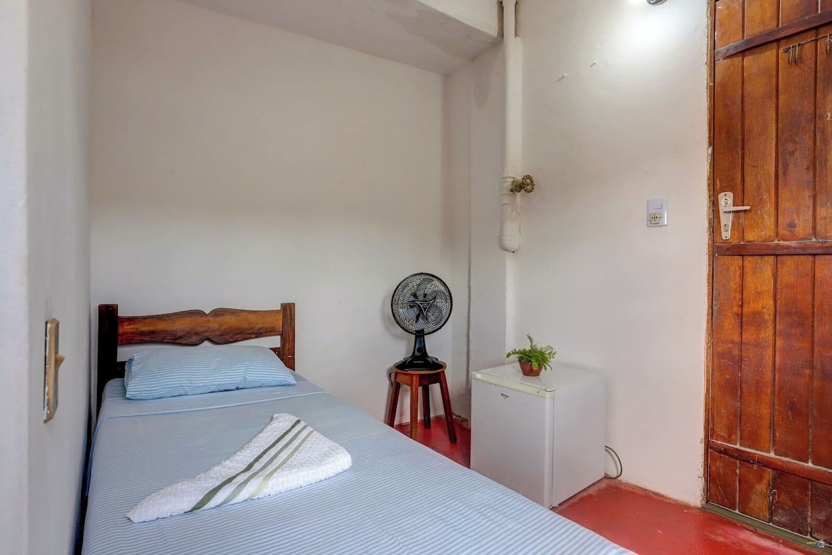 Une petite pièce étroite, avec un lit simple, une petite armoire blanche, un ventilateur et une porte en bois.