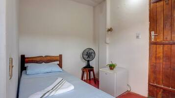 A small, narrow room with a single bed, a small white cabinet, a fan, and a wooden door.