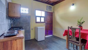 Una cocina con estufa a gas, refrigerador, mueble de madera, ventana con celosía decorativa y una mesa roja con una silla.