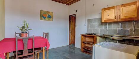 A room with a dining table, chairs, a potted plant, a kitchen area with cabinets, and a tiled floor.