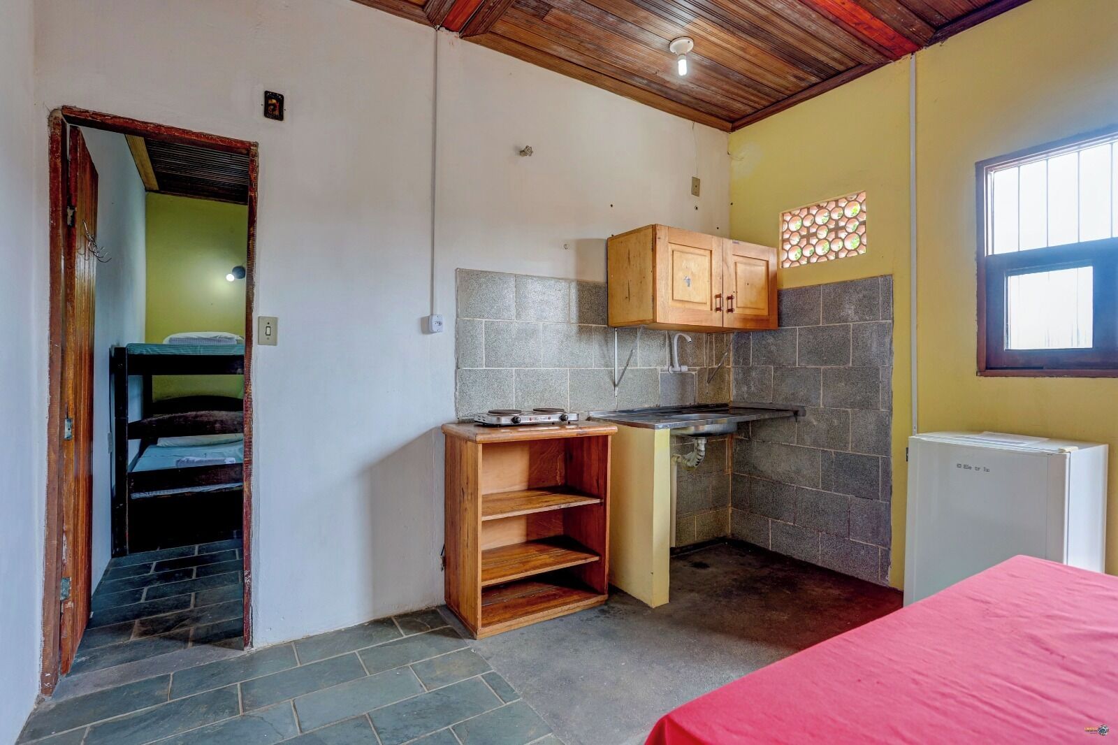 A compact kitchen with a sink, wooden cabinets, and a red bed.