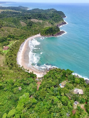 An aerial view of a coastal area with a beach, dense greenery, and a few buildings. - Pousada Pedra Solitária (Itacaré)