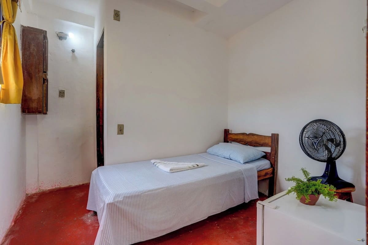 A small, single-bed room with a wooden headboard, a bedside fan, a potted plant, and a white cabinet.