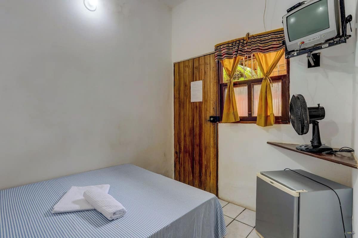 Une petite pièce avec un lit simple, un téléviseur fixé au mur, un ventilateur et une porte en bois.