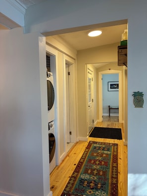 Een gang met een houten vloer, een kamerbreed tapijt, een wasmachine en een deur naar een andere kamer.