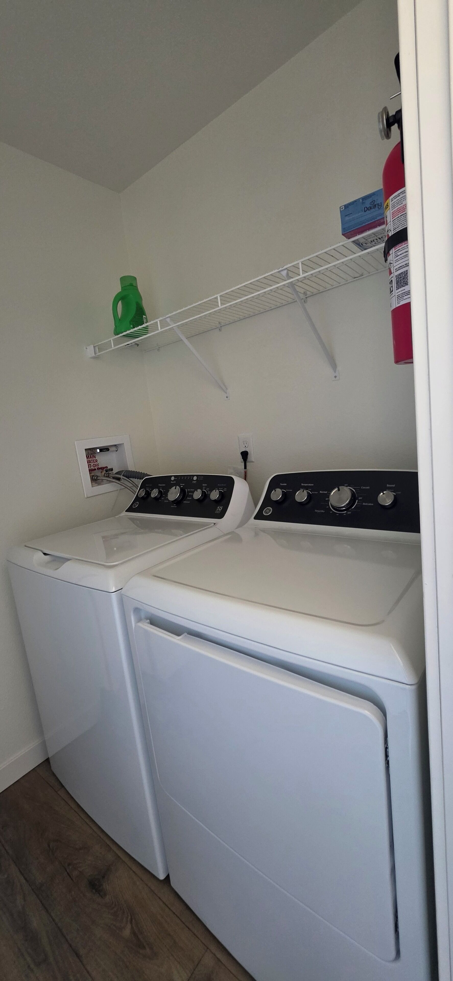 A laundry area with two white front-loading washers and dryers, a green water dispenser, and a white shelf with a box of detergent.