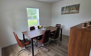 Une salle à manger avec une table en bois, quatre chaises, un vase de fleurs, un tableau au mur et une fenêtre avec des stores.