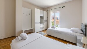 Une chambre avec deux lits, une armoire, un bureau et une fenêtre donnant sur des immeubles.