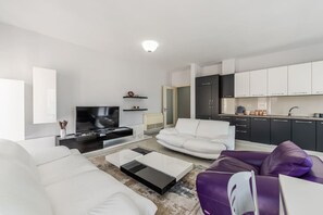Ein modernes Wohnzimmer mit einem Flachbildfernseher, weißen Sofas, einem Couchtisch und einem violetten Hocker.