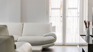 Un salon moderne avec un canapé blanc, une table basse et une grande fenêtre ornée de rideaux légers.