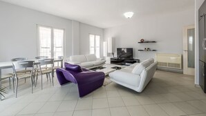 Un salon moderne comprenant un coin repas, des canapés blancs, un téléviseur à écran plat et un radiateur encastré.