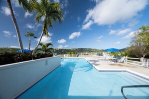 Une piscine bordée de palmiers, une clôture blanche et une vue sur des collines lointaines.