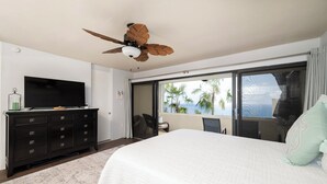 A bedroom with a bed, a television on a dresser, a ceiling fan, and a large window with a view of palm trees. - Brand New Centrally Located  2BD/1BA Ocean View (St. Thomas)