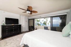 A bedroom with a bed, a television on a dresser, a ceiling fan, and a large window with a view of palm trees. - Brand New Centrally Located  2BD/1BA Ocean View (St. Thomas)