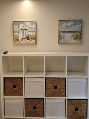 Une pièce décorée avec deux tableaux sur le thème de la plage, une étagère blanche avec des paniers en osier, et une télécommande posée sur l’étagère.