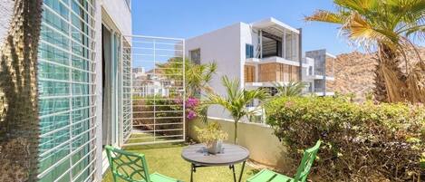 Un edificio moderno con un balcón, sillas verdes, una mesita y una planta en maceta.