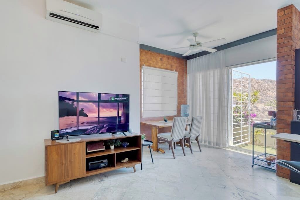 Un salon moderne doté d’un téléviseur à écran plat, d’un meuble de rangement en bois, d’un coin repas avec des chaises et d’une grande porte-fenêtre coulissante donnant sur un balcon.