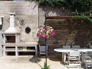 An outdoor patio with a stone wall, a grill, a round table with chairs, and a hanging flower basket. - Well-equipped 8-person house with very large swimming pool. very close to town center. (Nîmes)