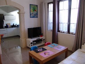 A living room with a TV, a table with children's placemats, a kitchen in the background, and a window with curtains. - Well-equipped 8-person house with very large swimming pool. very close to town center. (Nîmes)