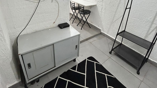 Una habitación compacta con televisión montada en la pared, una pequeña mesa con sillas, un perchero metálico y un armario.