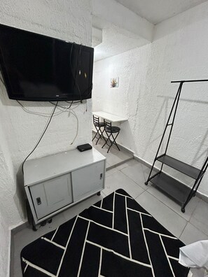Une petite pièce, de taille modeste, avec un téléviseur à écran plat fixé au mur, un bureau blanc avec une chaise, une petite armoire, un tapis à motifs noir et blanc et une étagère en métal.