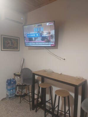Ein Zimmer mit an der Wand montiertem Fernseher, einem Holztisch mit zwei Hocker, einem Wasserspender und einem gerahmten Bild.