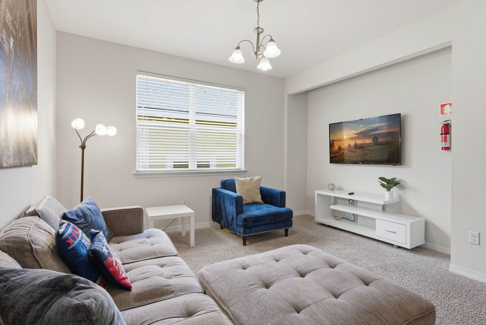 Uma sala de estar com sofá, banquinho, poltrona azul, rack branco para TV e um quadro na parede.