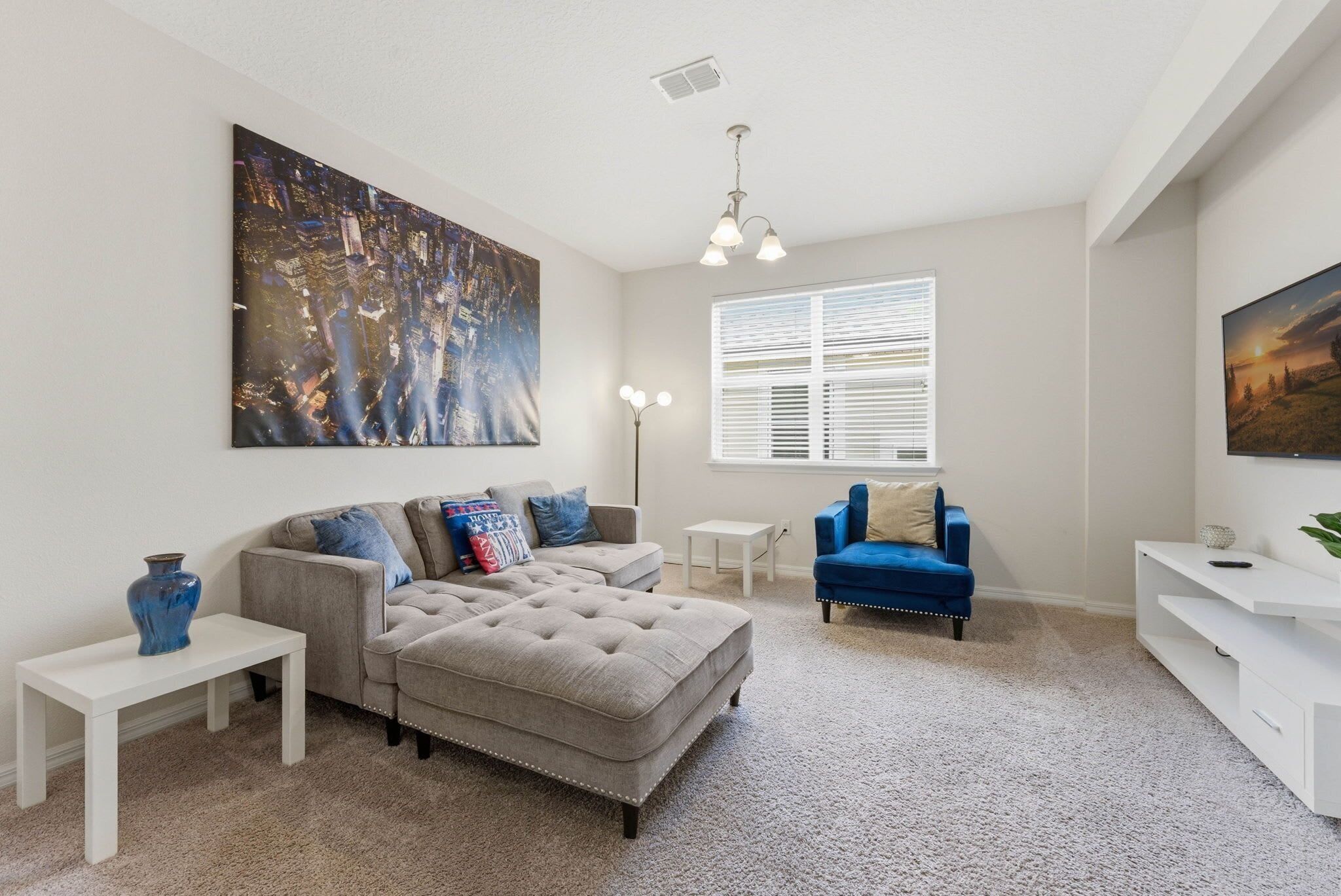 A living room with a grey sectional sofa, a blue armchair, a white coffee table, a white side table, and a large painting on the wall.