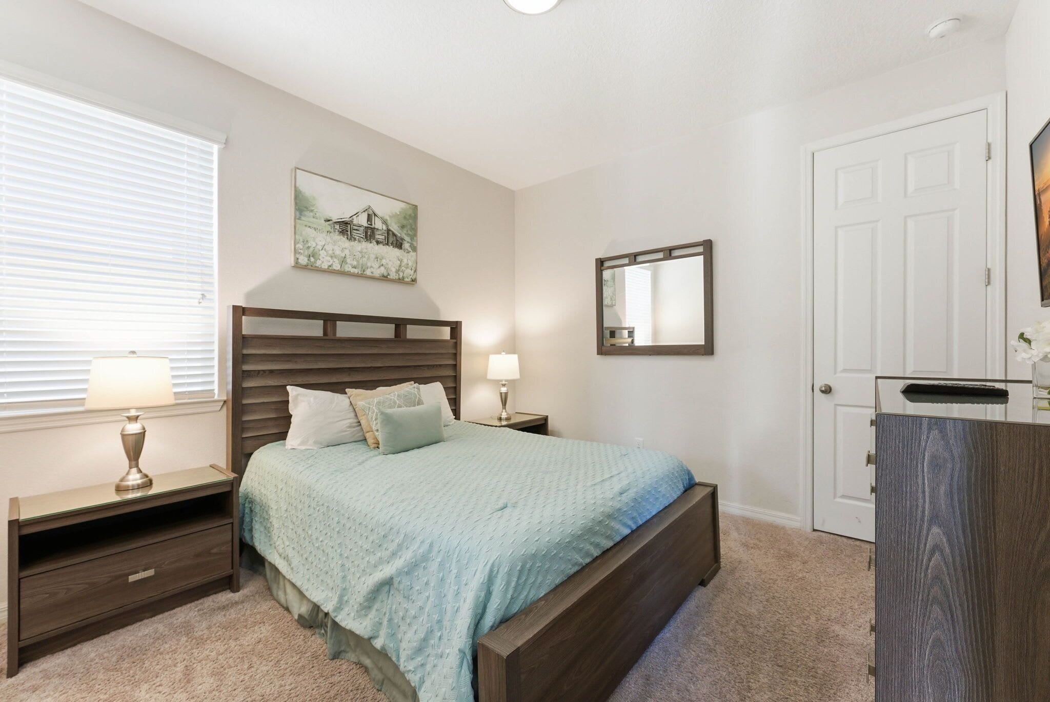 A bedroom with a bed, nightstands, a dresser, a window with blinds, a painting, and a door.