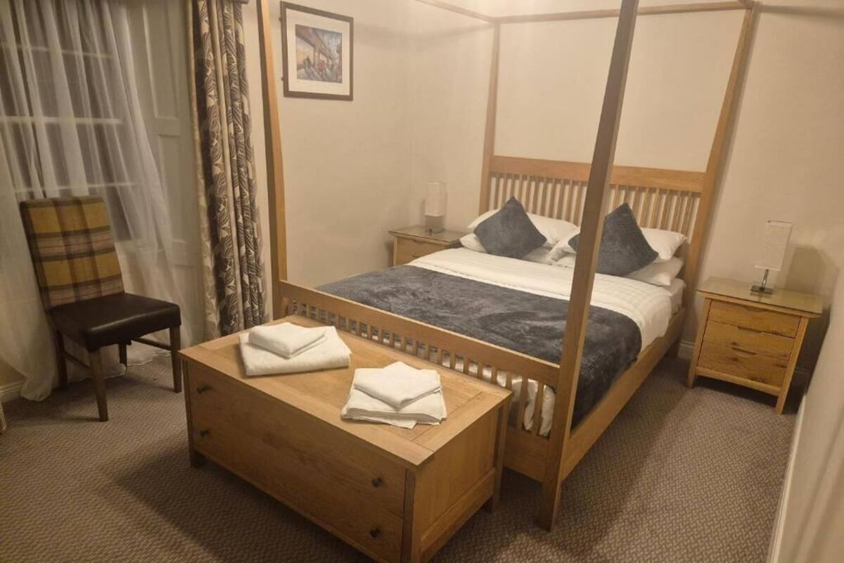 En säng med fyra stänger och en träram, ett sängbord med en lampa och en stol med en rutig kudde.