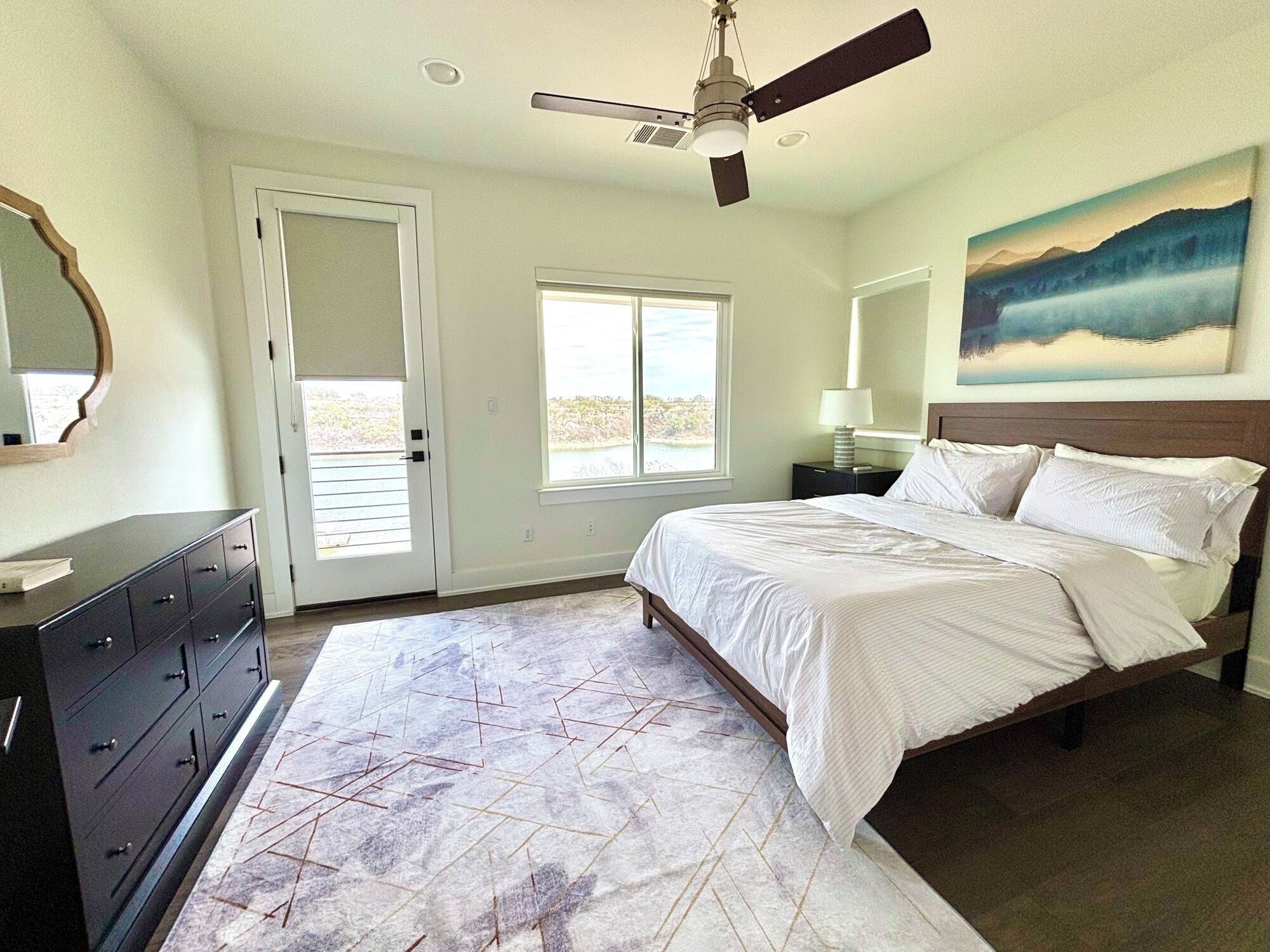A bedroom with a bed, a ceiling fan, a door, a window, a nightstand, and a painting on the wall.