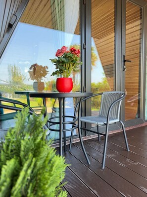 En modern utomhusplats med sittmöbler, ett glasbord och stolar, en röd vas med blommor och utsikt över grönskan genom glaset.