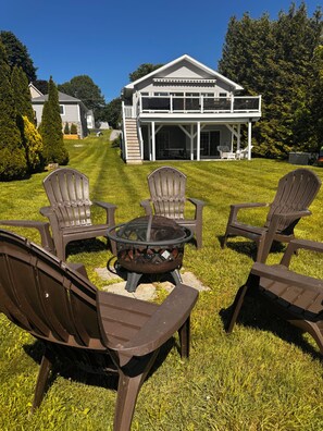 En uteplats med fyra Adirondack-stolar och en eldstad, med ett tvåvåningshus med en täckt veranda som bakgrund.