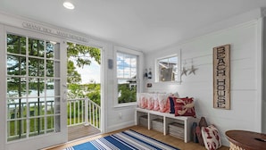 Una habitación con un gran ventanal corredero, un banco con almohadas rojas y blancas, un banco de madera, un letrero en la pared que dice 'BEACH HOUSE' y una alfombra azul a rayas.