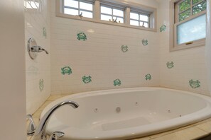 Un baño con una bañera independiente, paredes cubiertas de azulejos blancos con detalles decorativos verdes, un ventanal sobre la bañera y un cabezal de ducha visible.