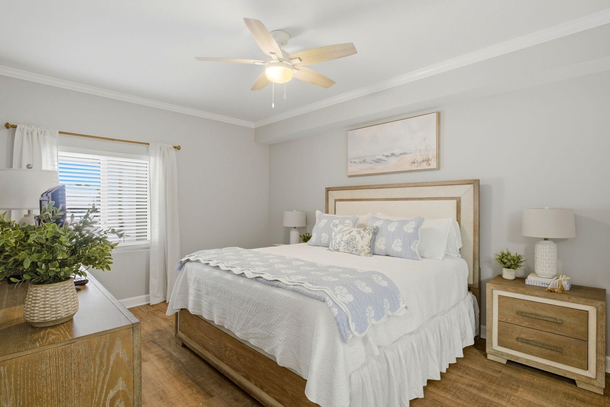 A bedroom with a bed, bedside tables, a ceiling fan, a window with blinds, and a wall-mounted painting.