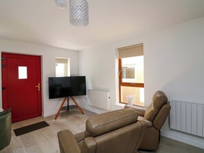 A living room with a red door, a flat-screen TV on a stand, a brown leather sofa, a radiator, and a window with blinds. - The Mews @ Suskar (Glendalough)