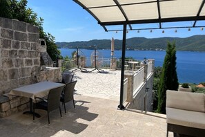 Een terras met een tafel en stoelen, een stenen muur en uitzicht op een meer en heuvels.