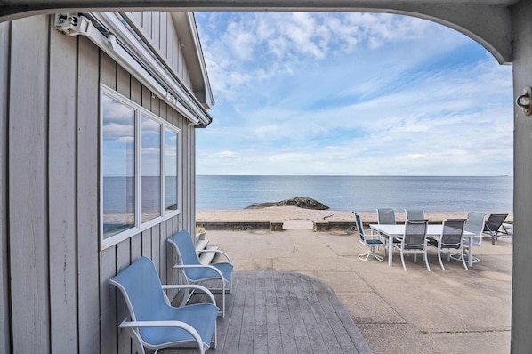 A covered outdoor seating area with a view of the sea, a concrete deck, and a building with large windows.